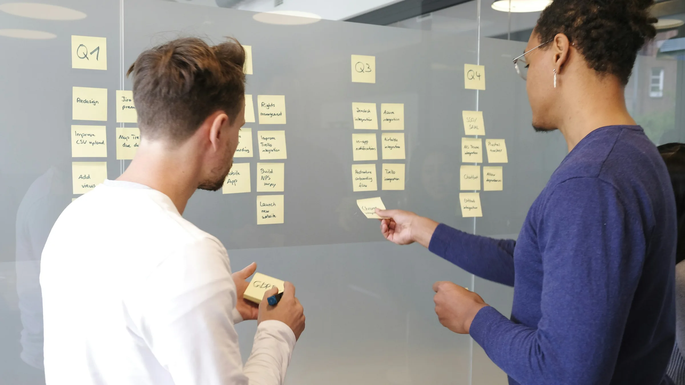 Two people arranging sticky notes into columns on a glass wall
